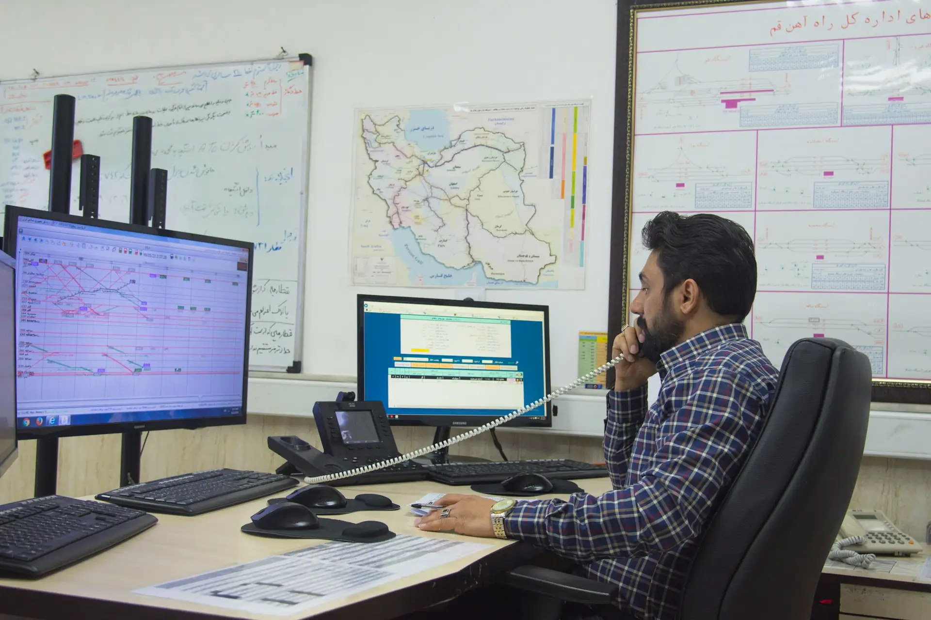 A coordinator on the phone at a desk with two monitors, maps and planning boards on the wall behind him