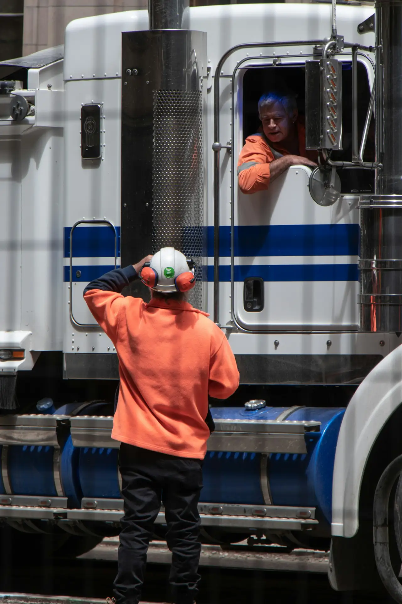 A yard worker in a high-visibility top and ear defenders talking with a truck driver leaning from his cab