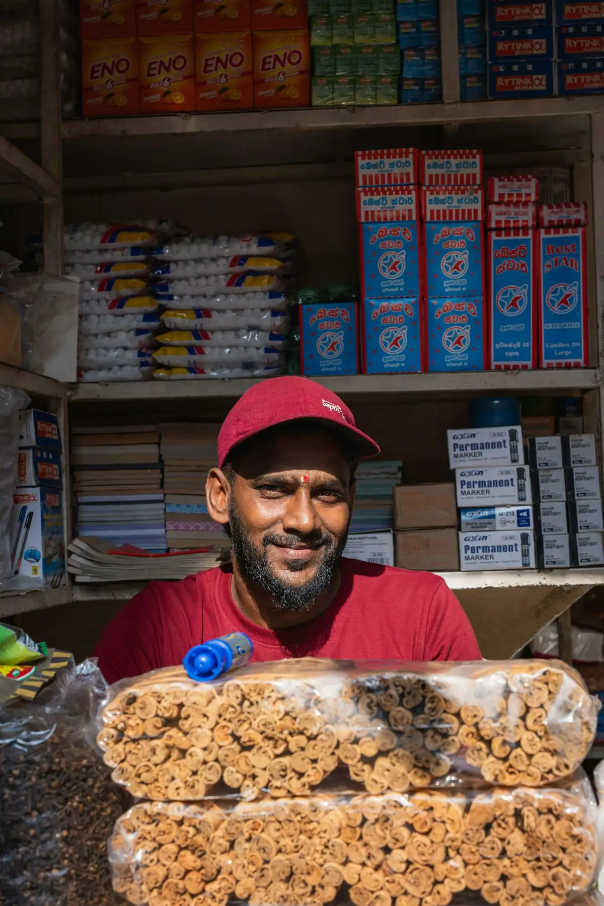 A shopkeeper in a red cap smiling in front of stocked shelves